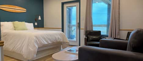 A hotel room with a bed, a round table, a sofa, a chair, a potted plant, and a view of the sea through a window.