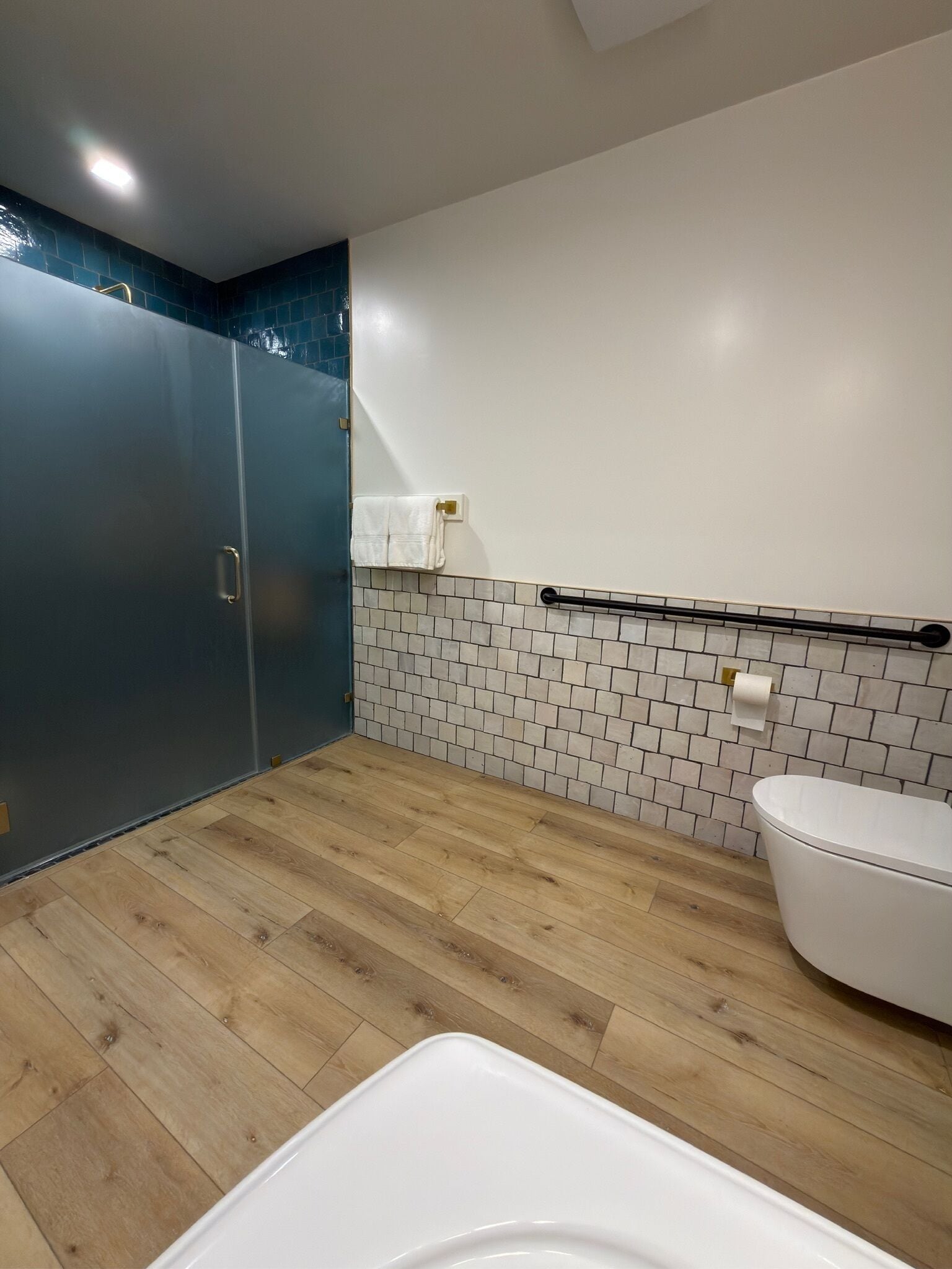 Un baño moderno con piso de madera, inodoro blanco, toallero y zona de ducha con pared de azulejos azul oscuro.