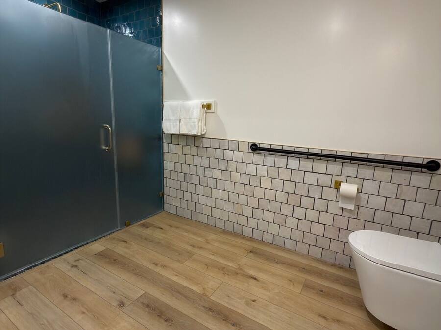 Un baño moderno con piso de madera, inodoro blanco, toallero y zona de ducha con pared de azulejos azul oscuro.