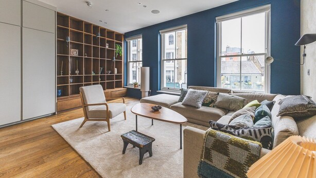 Una sala de estar moderna con sofá, sillón y mesa de centro, una estantería y ventanas amplias con vista a la ciudad.