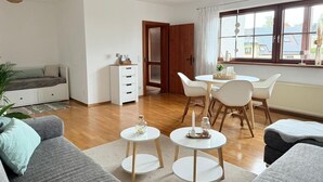 A modern living room with a sofa, a dining table with chairs, a small white cabinet, and a large window with curtains. - NEW! Time out at the Hirzenkopf (Steinberg-Deckenhardt)