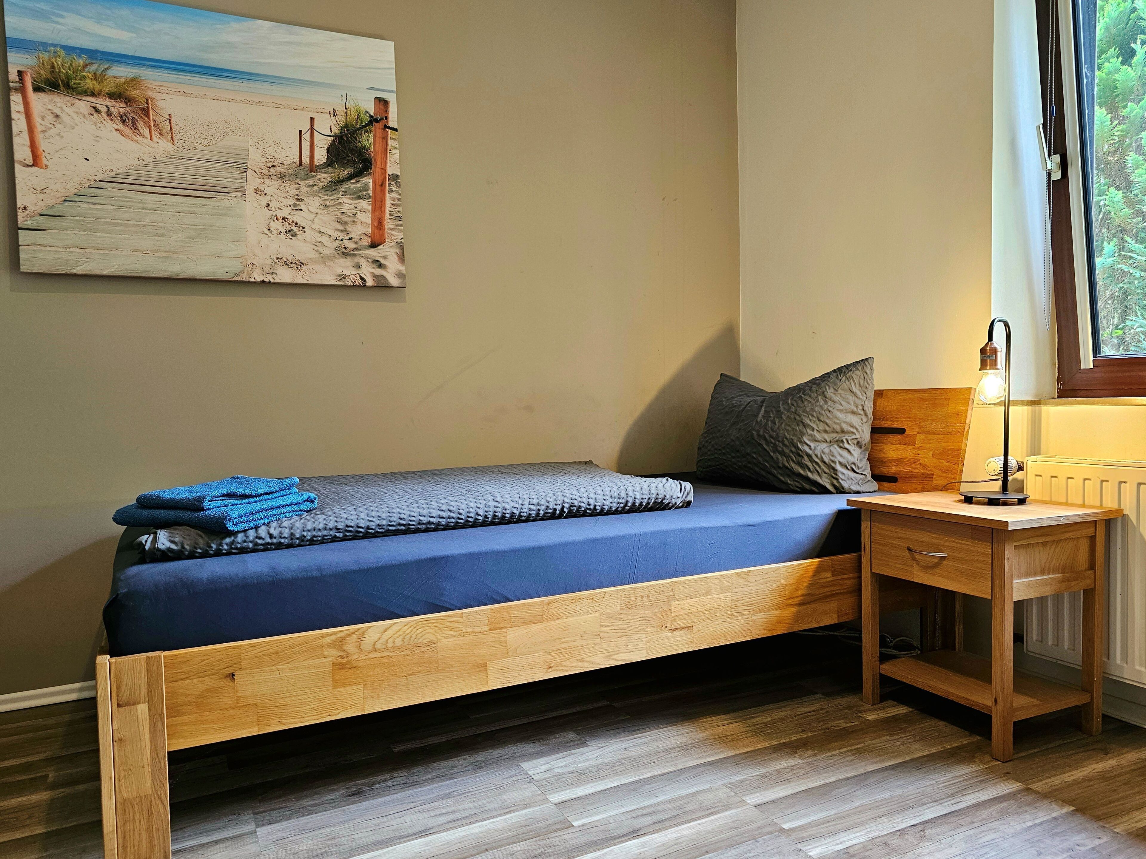 Una cama individual con estructura de madera y colchón azul, una mesita de noche con lámpara, y una ventana con vistas a zonas verdes.