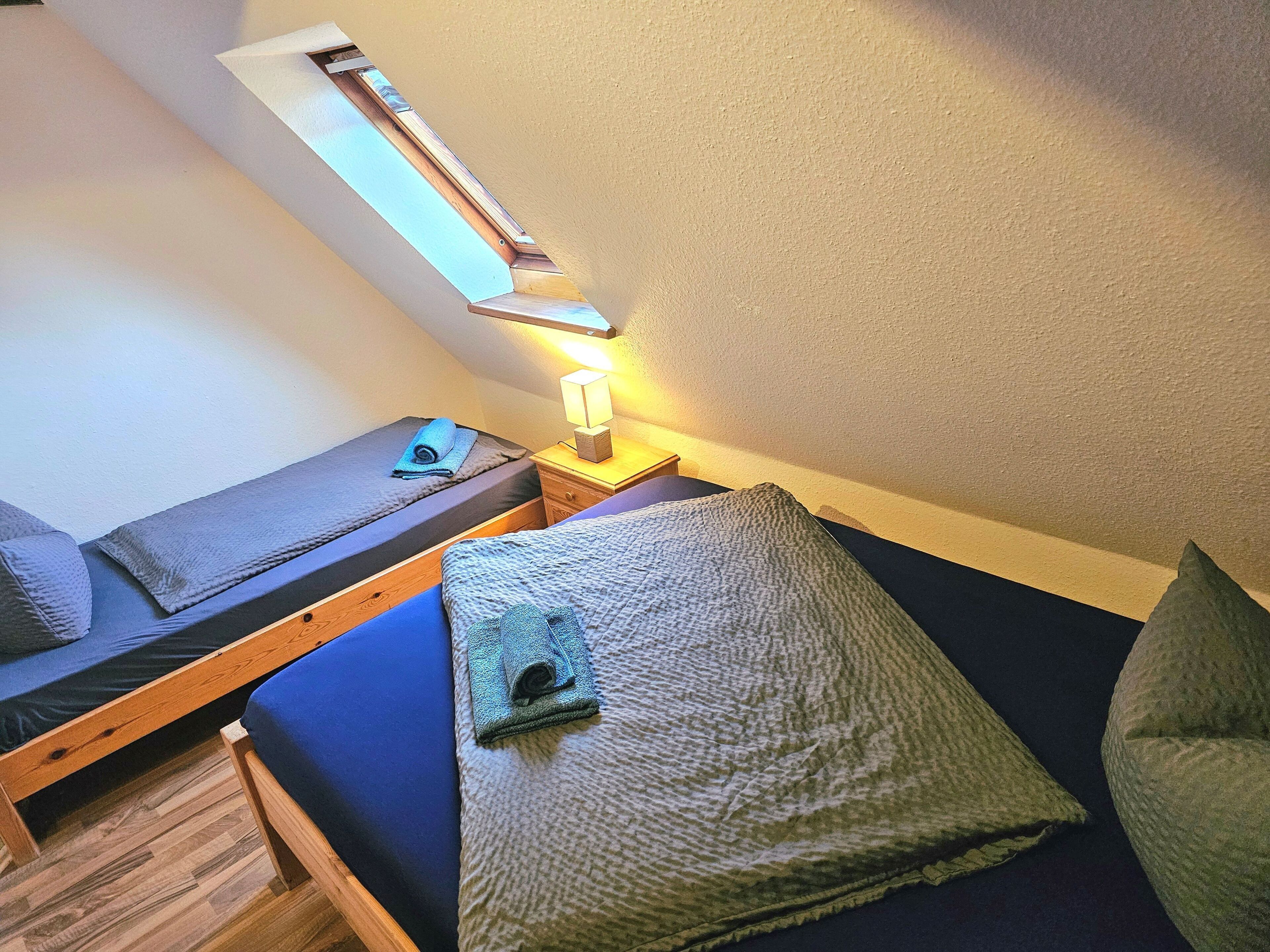 Une petite chambre avec un lit simple, une lucarne, une table de chevet avec une lampe et un plancher en bois.