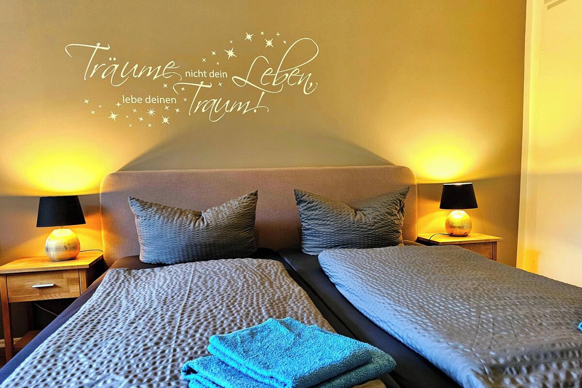 Ein Doppelbett mit Kopfteil, zwei Nachttische mit Lampen und eine Wand mit dekorativem Spruch.