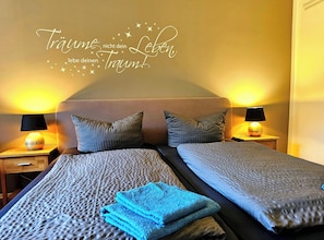 Un lit double avec une tête de lit, deux oreillers, une table de chevet avec une lampe et un mur orné d’une inscription décorative.
