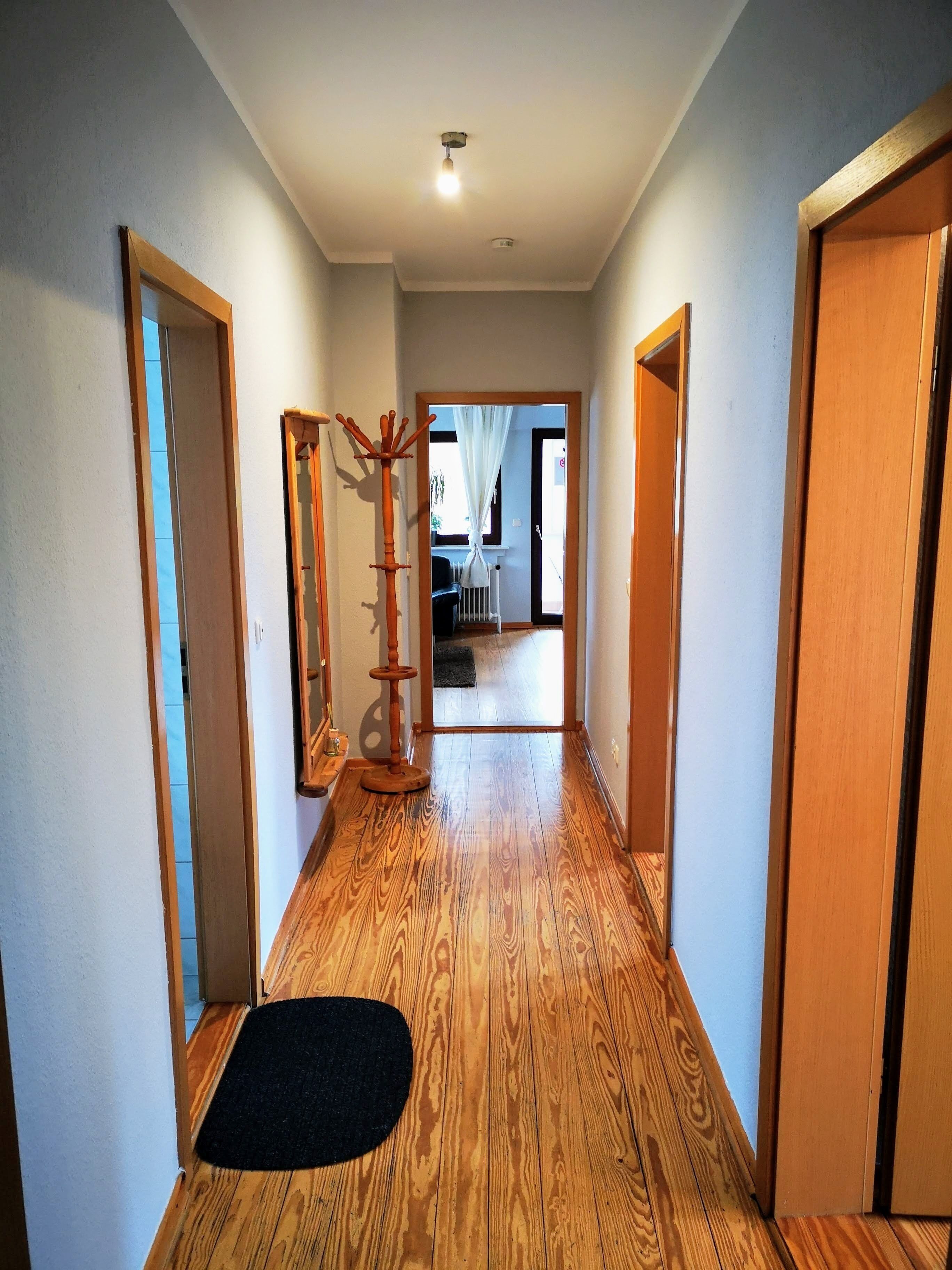 Un couloir doté d’un plancher en bois, d’un paillasson noir, d’un porte-manteau et d’une porte donnant sur une autre pièce.