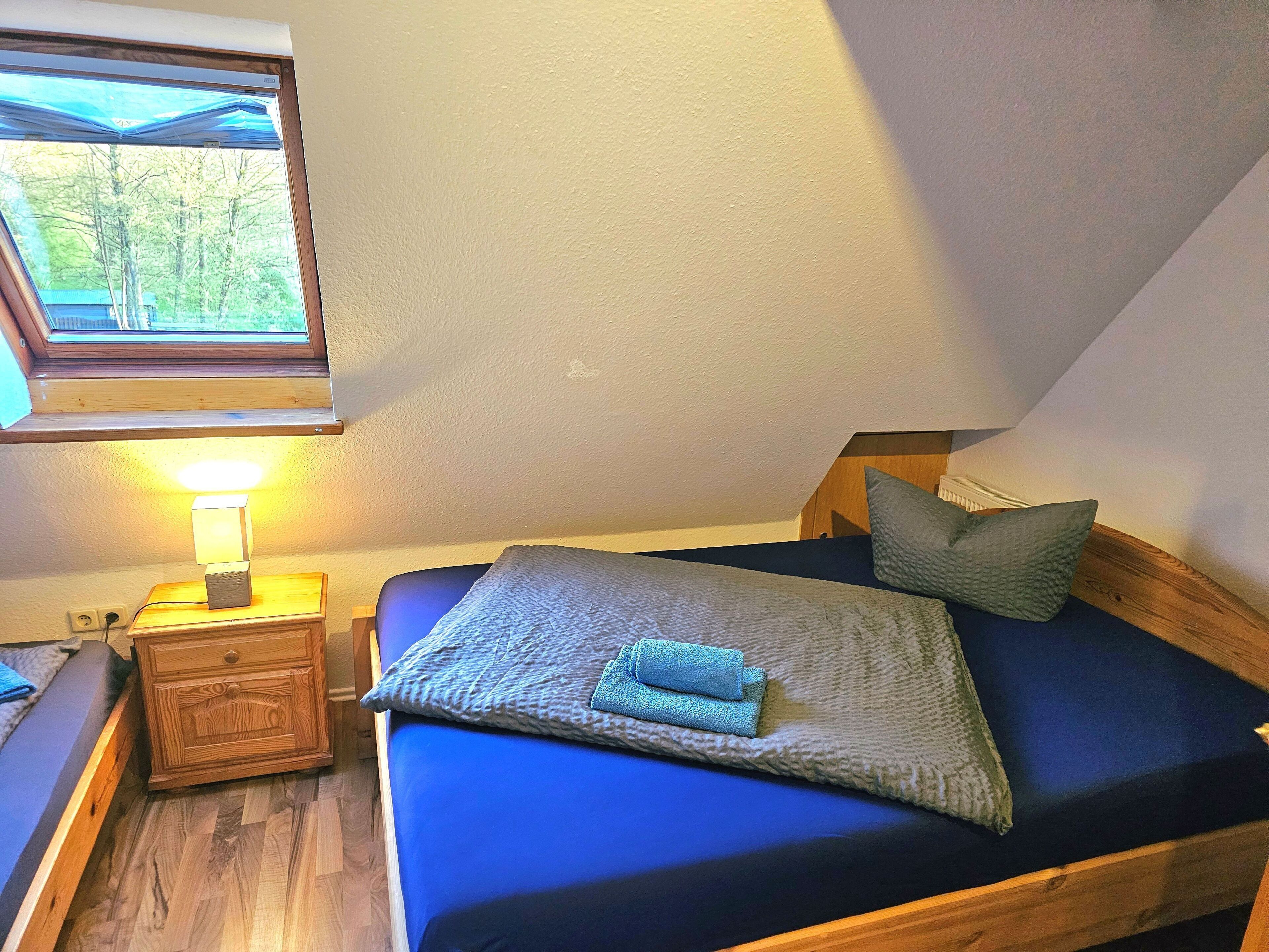 A bedroom with a bed, a nightstand with a lamp, a window, and a view of trees.