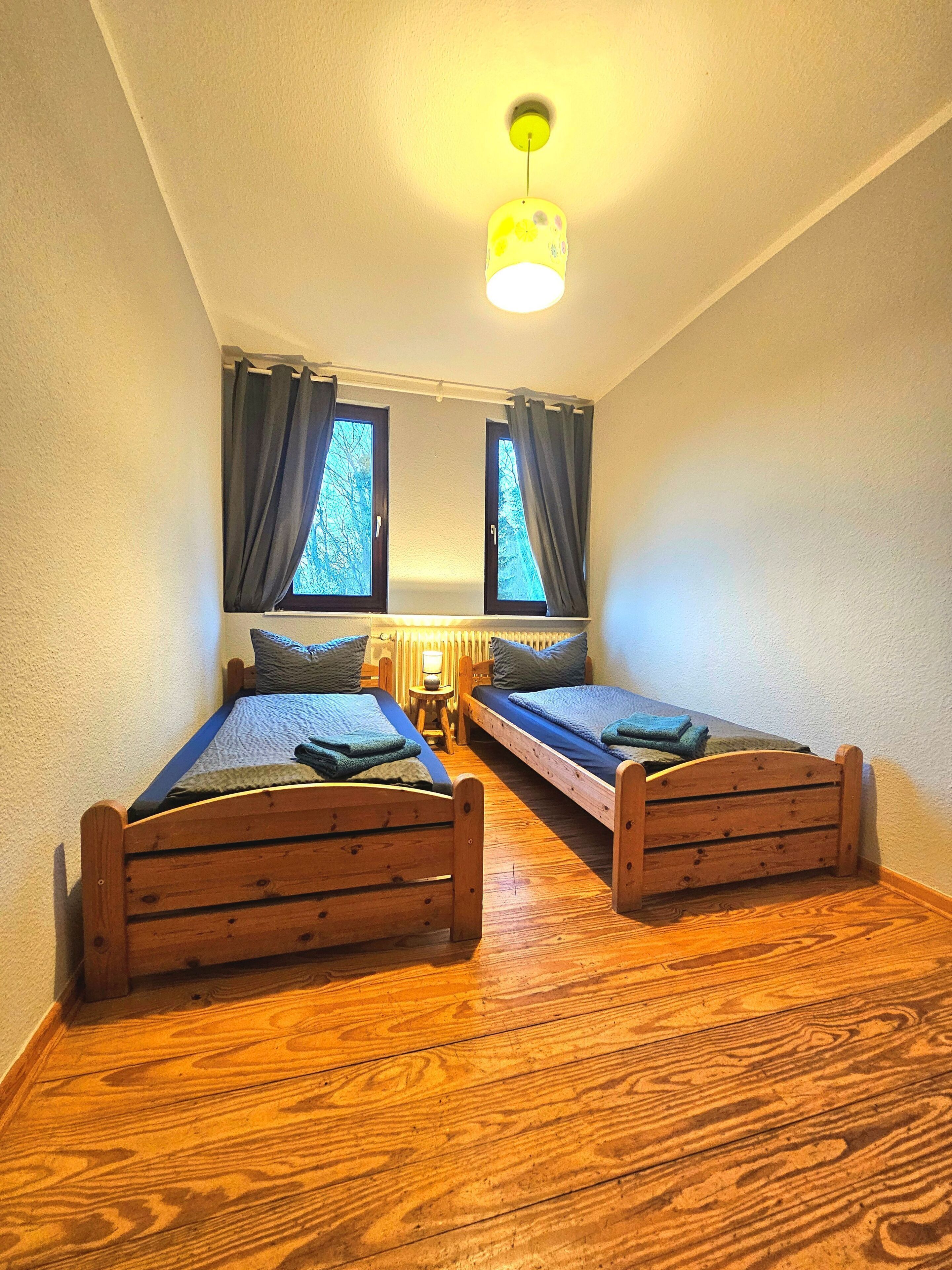 A room with two wooden beds, a small table, a lamp, and a ceiling light fixture.