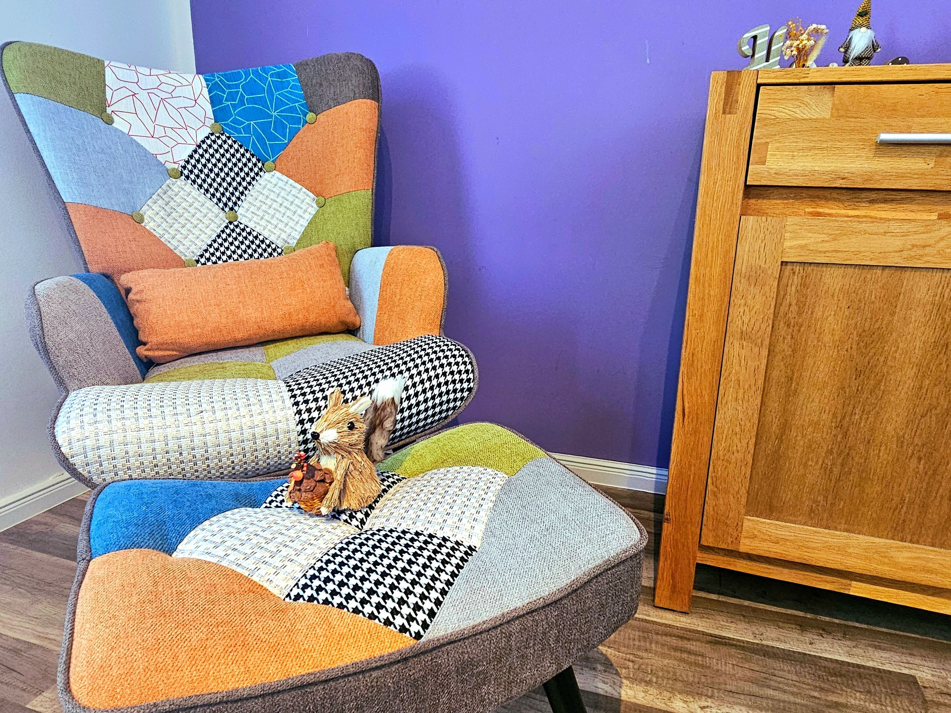 A colorful armchair with a patchwork design, a small figurine, and a wooden cabinet against a purple wall.