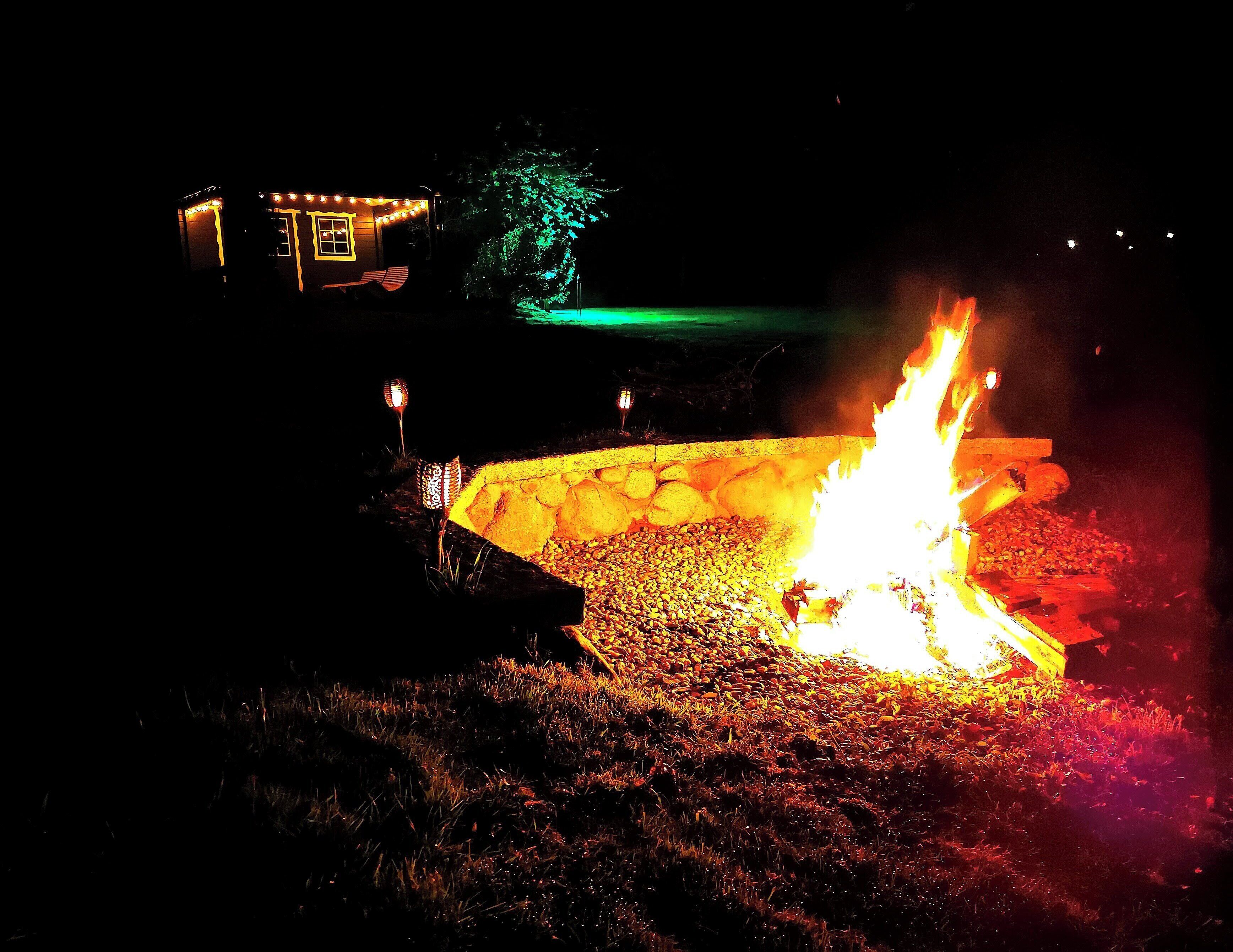 Escena nocturna al aire libre con un brasero rodeado de rocas y una estructura pequeña iluminada al fondo.