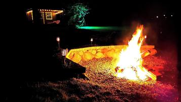 Escena nocturna al aire libre con un brasero rodeado de rocas y una estructura pequeña iluminada al fondo.