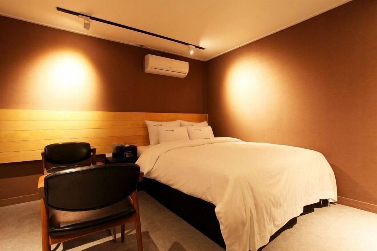 A hotel room with a bed, a chair, a desk, and an air conditioning unit.