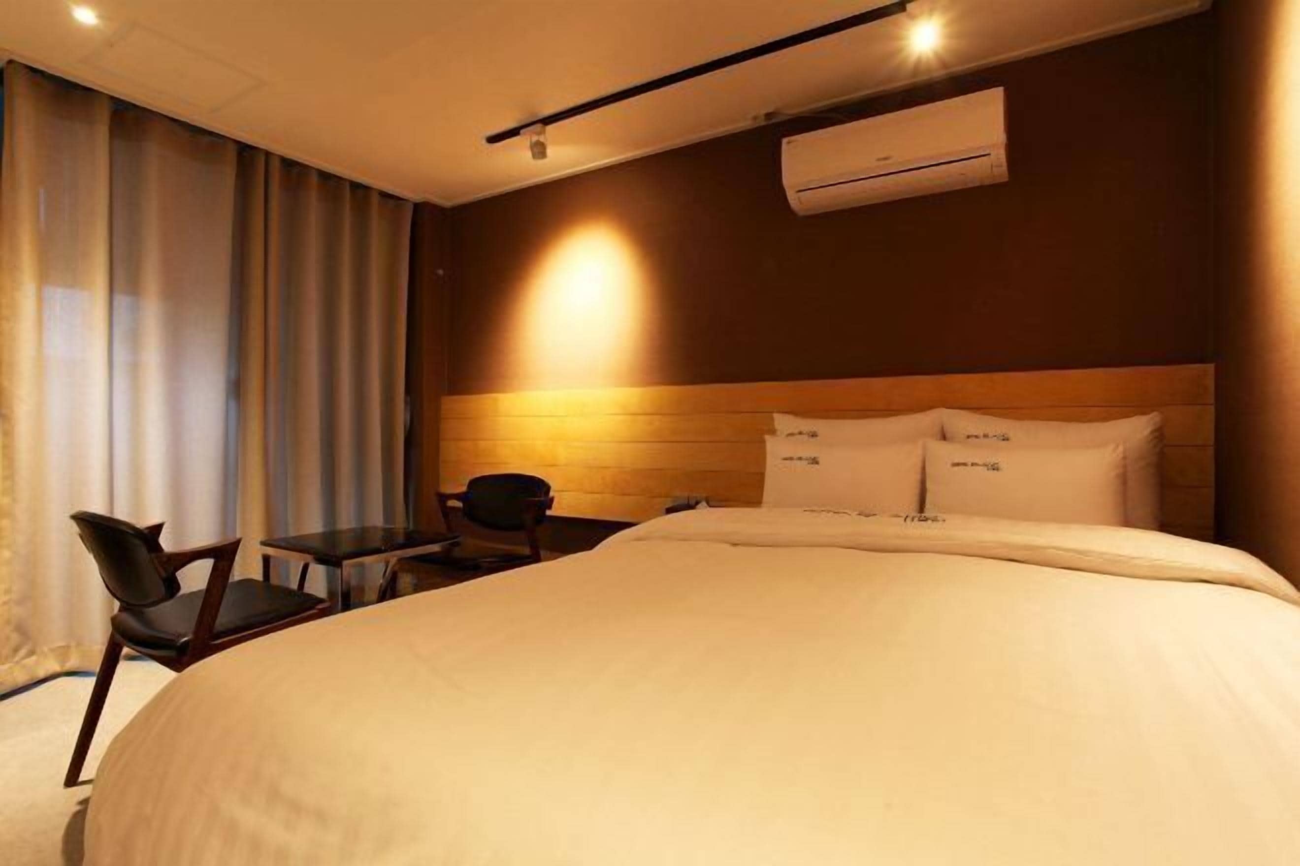 A hotel room with a large bed, a desk, two chairs, and an air conditioning unit on the wall.