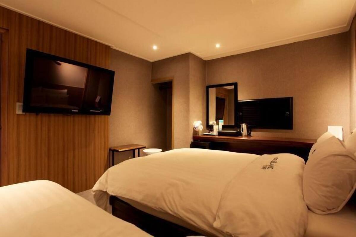 A hotel room with a bed, a TV mounted on the wall, a mirror, and a small table with a lamp.