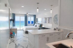 A modern hotel room with a large bed, a sitting area, a kitchen area, and a view of the ocean.