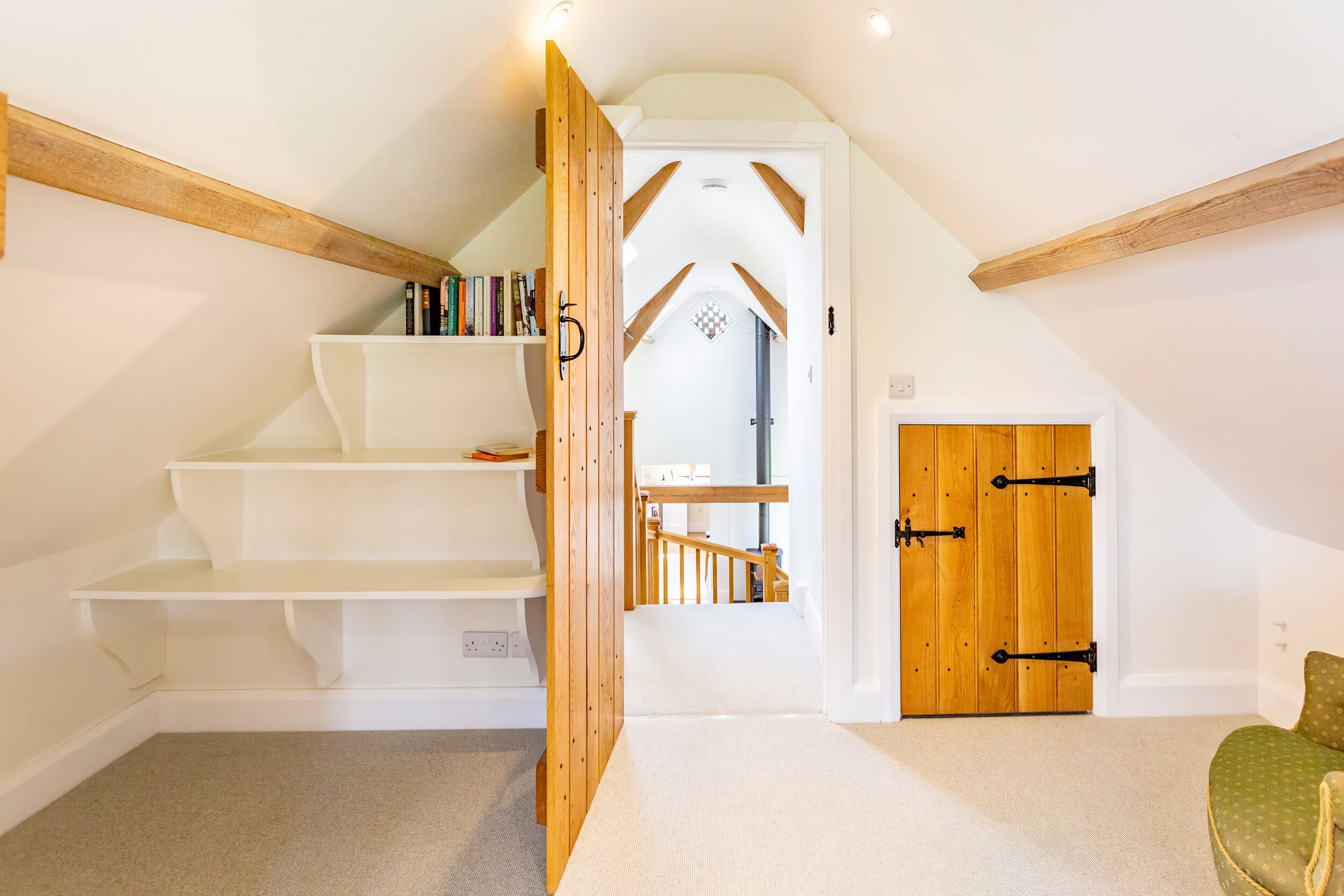 Un dormitorio con techo inclinado, vigas de madera, estantería empotrada, puerta de madera y sillón tapizado en verde.