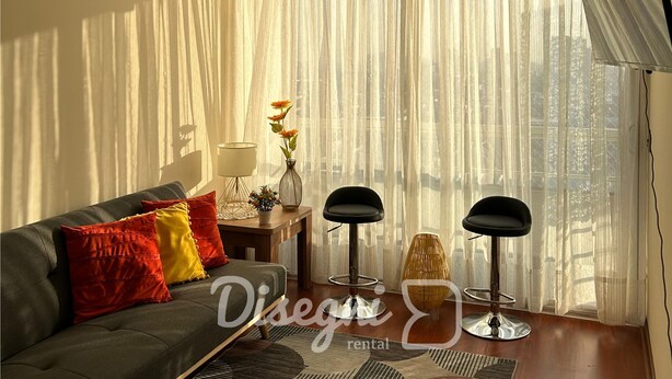 A modern living room with a grey sofa, red and yellow cushions, a small table with a vase of flowers, two black chairs, and sheer curtains. - Disegni 17 - Santiago Apartment, Close to Metro and Culture (Santiago)