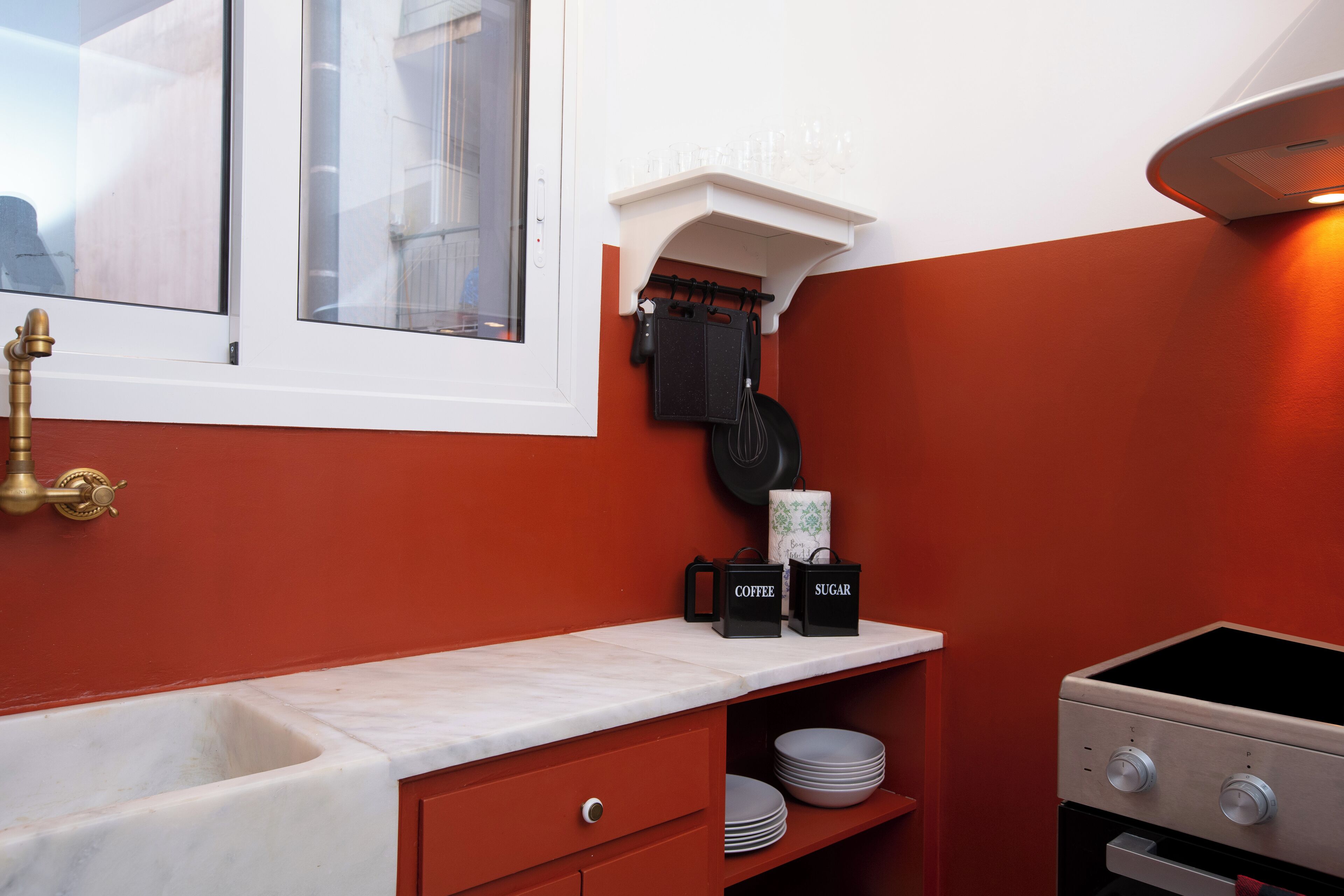 A kitchen with a white countertop, a red wall, a window, a sink, a stove, and a shelf with kitchen utensils.