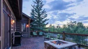 Una terraza de madera con un pozo de fuego de piedra, mobiliario exterior y vistas a árboles y al cielo.