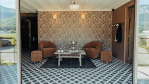 A room with two brown armchairs, a small table with a vase, patterned wallpaper, and a view of a car and greenery outside.