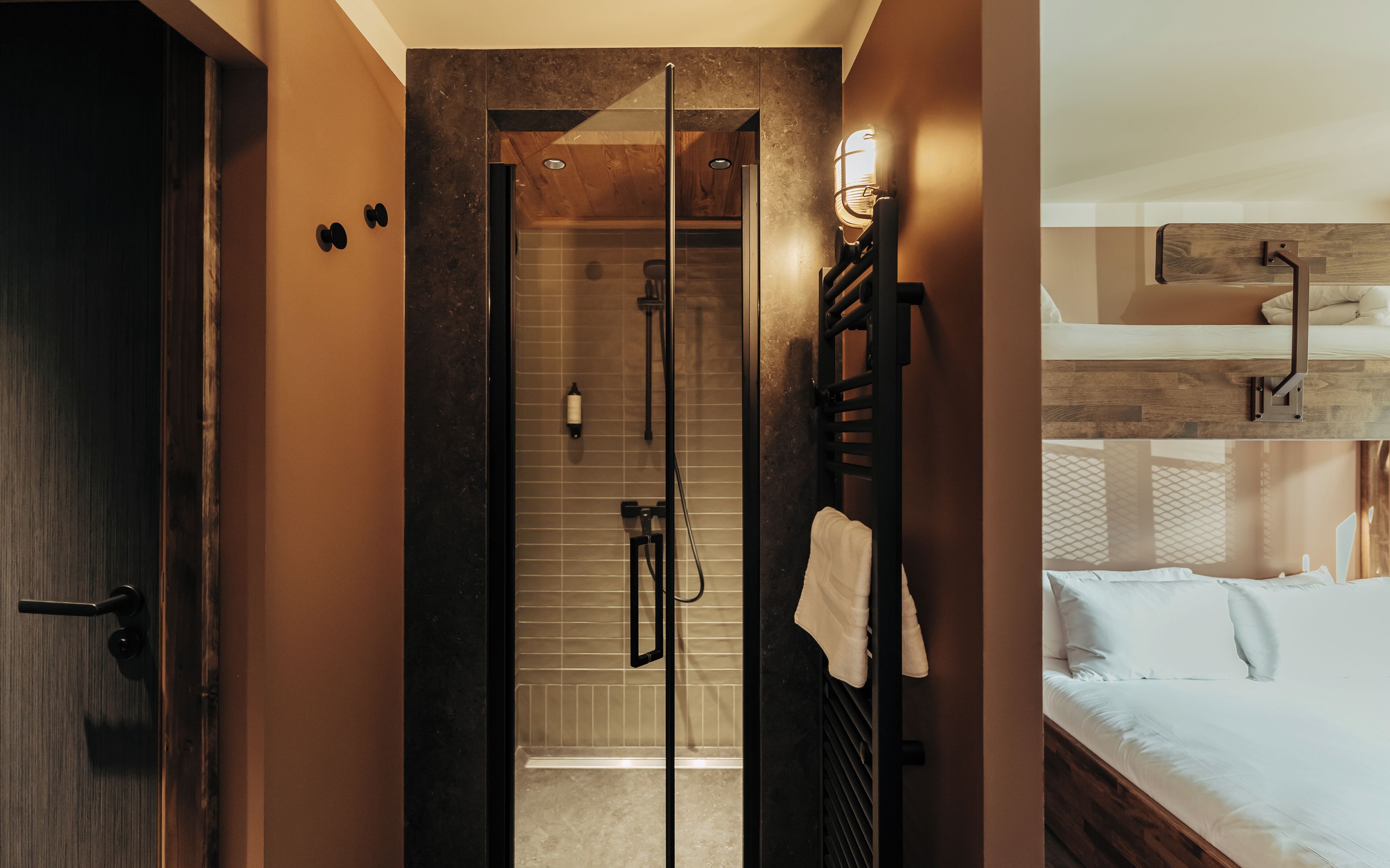 Una habitación de hotel moderna con puerta de madera, ducha con cerramiento de vidrio, una toalla colgada en un perchero y una cama con cabecero de madera.