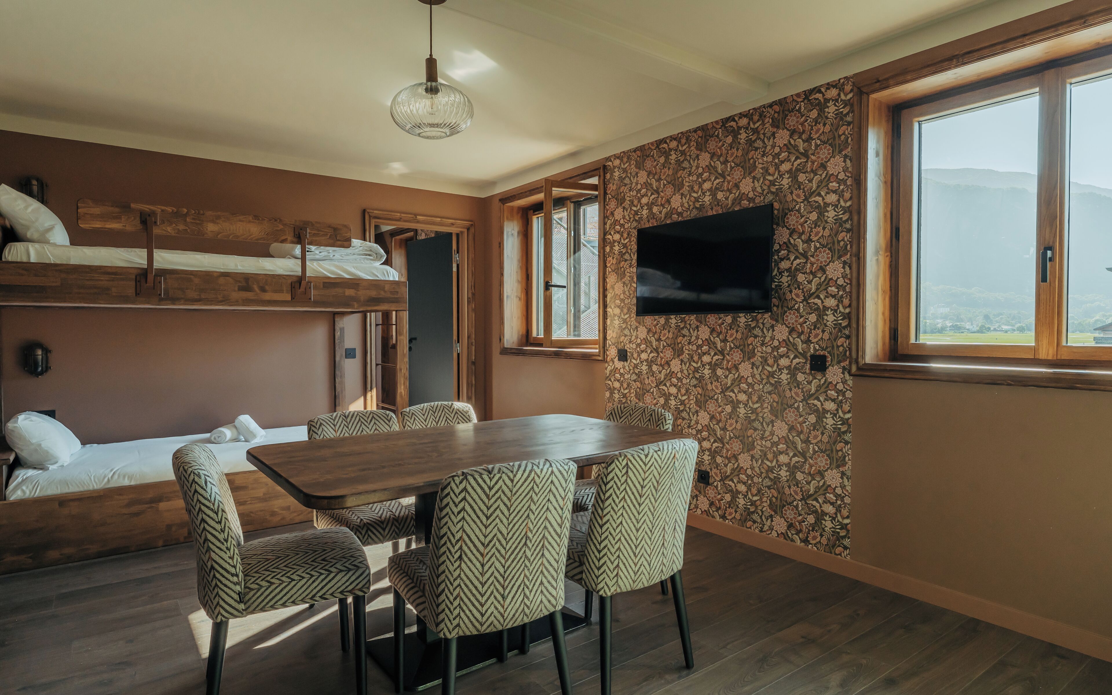 Habitación pequeña con mesa de madera, cuatro sillas estampadas, una cama individual alta, televisor y ventana grande con vistas.