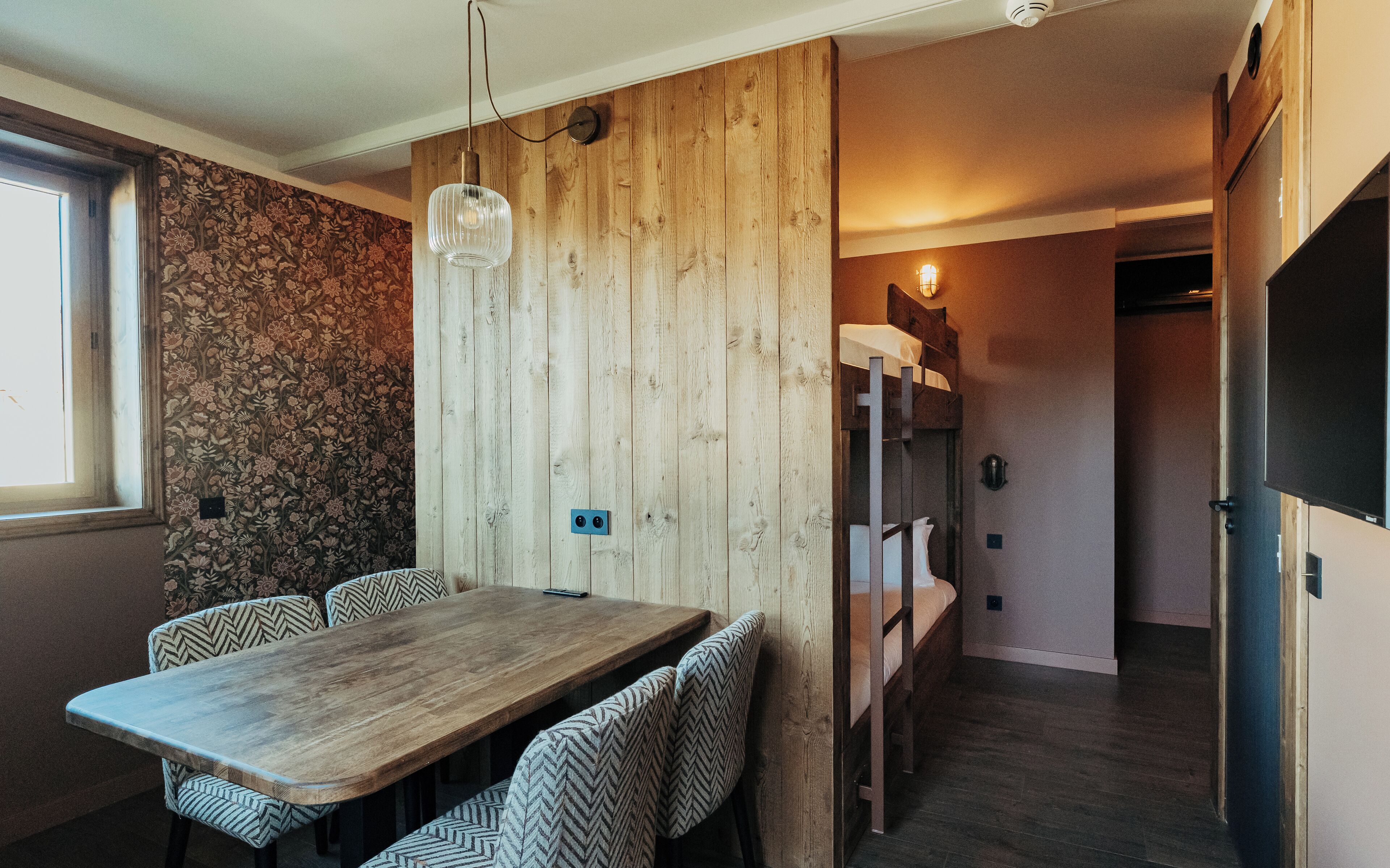 Un comedor con mesa de madera y sillas estampadas, una pared de madera con un panel de papel tapiz floral, y una cama litera al fondo.