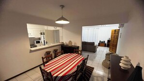 Un coin repas avec une nappe à rayures, des chaises en bois, un luminaire suspendu, une cuisine en arrière-plan et un salon avec un canapé et des rideaux.