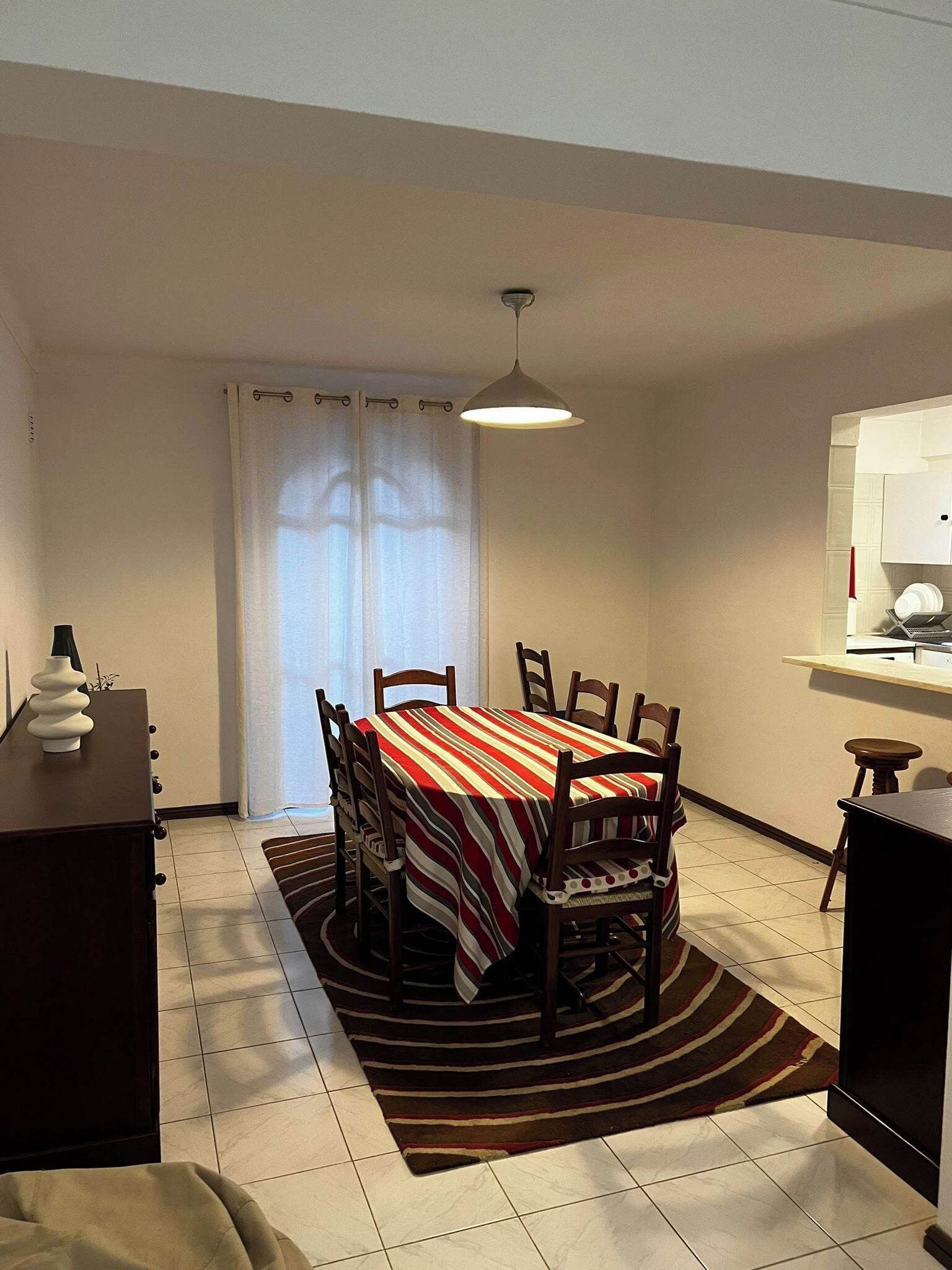 A dining area with a table set for six, a ceiling light, a window with curtains, and a small kitchen area with a microwave.