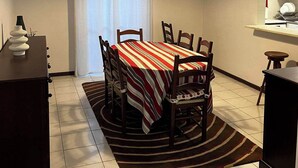 Un coin repas avec une table dressée pour six personnes, un luminaire au plafond, une fenêtre avec des rideaux et un petit espace cuisine équipé d’un four à micro-ondes.