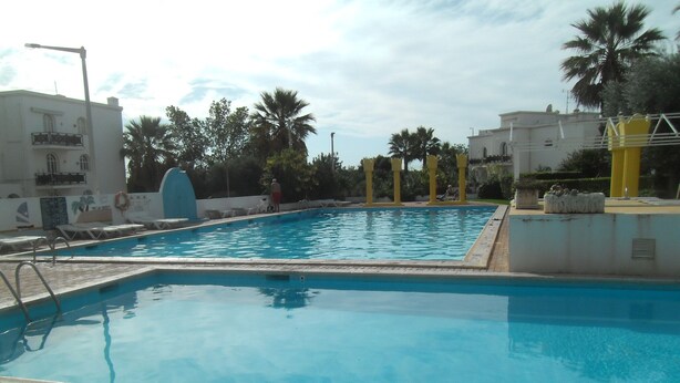 Une piscine dotée d’une terrasse carrelée, entourée de palmiers et de bâtiments.