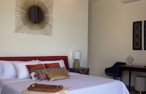 Una cama bien hecha con cabecera roja, un espejo con forma de sol montado en la pared, un escritorio con silla y una mesita de noche con lámpara.