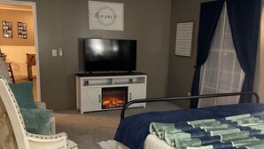 A bedroom with a bed, a TV, a fireplace, a chair, and a ceiling fan.