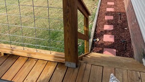 A wooden deck with a metal railing, a brick pathway, and a view of a grassy yard and trees.
