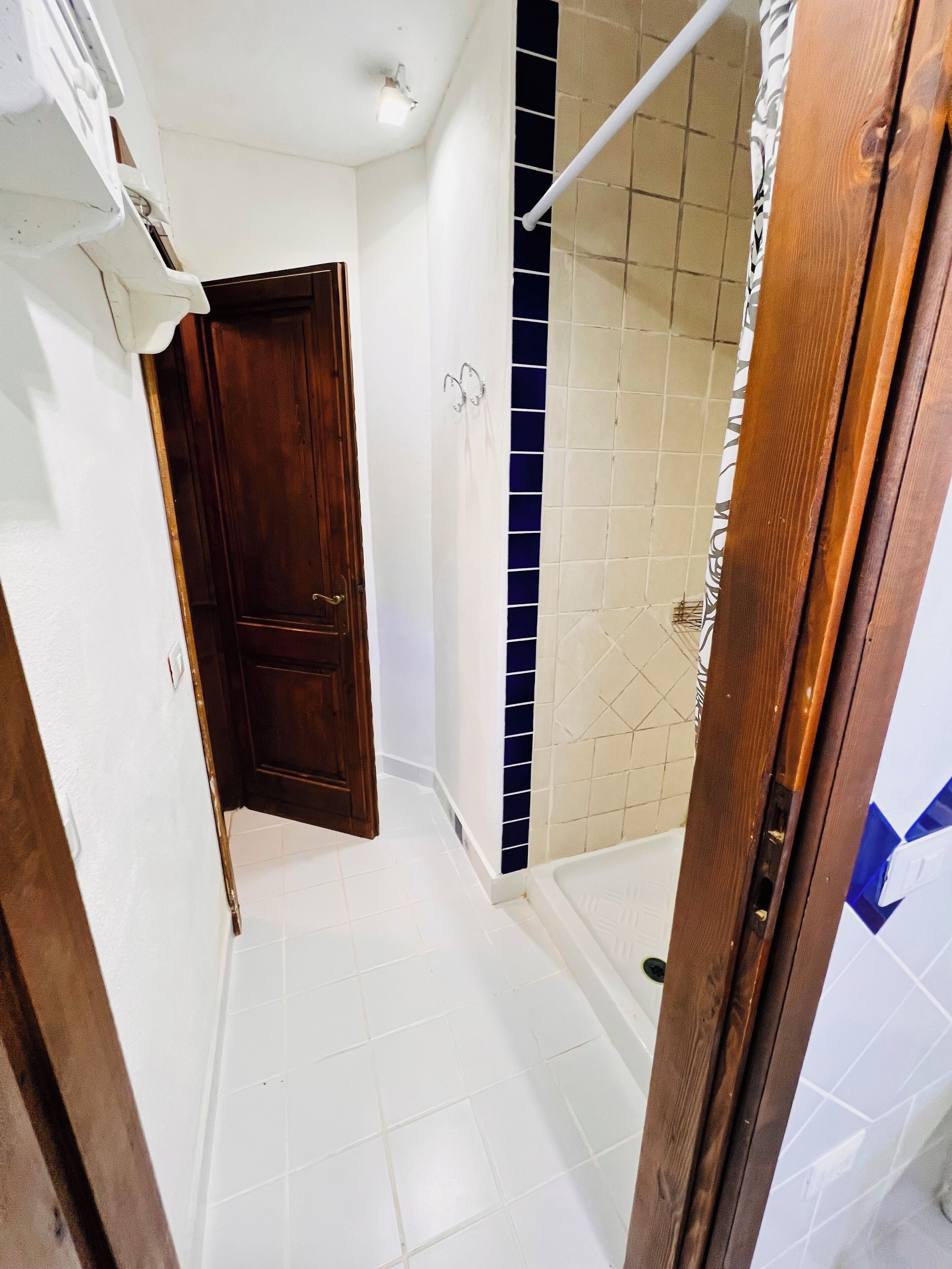 Ένα μικρό μπάνιο με λευκά πλακάκια στο δάπεδο, μια ξύλινη πόρτα και μια περιοχή με ντους, με πλακάκια σε μπεζ και μπλε αποχρώσεις.