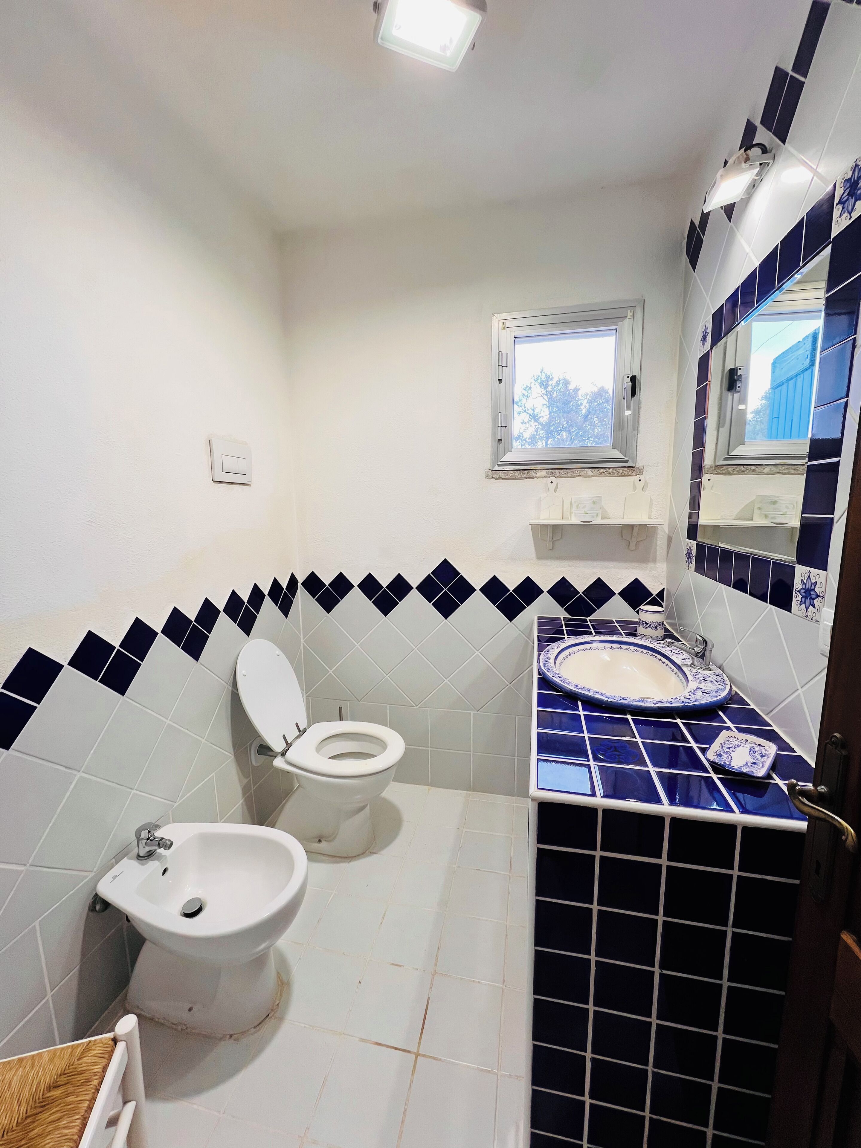 A bathroom with two toilets, a bidet, a sink with a blue-tiled vanity, and a window above the sink.