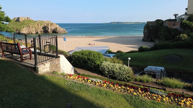 A coastal view with a beach, a flag, a bench, and a garden with flowers. - Sea Haze (Tenby)