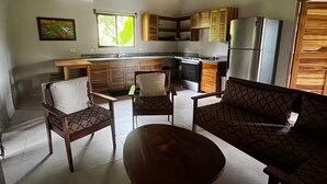 A modern kitchen with wooden cabinets, a built-in stove, and a refrigerator. There is a dining area with a round table and two chairs. - Cool Comfort & Local Charm (Savegre de Aguirre)