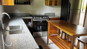 A kitchen with granite countertops, wooden cabinets, a stove, a refrigerator, and a wooden dining table. - Cool Comfort & Local Charm (Savegre de Aguirre)