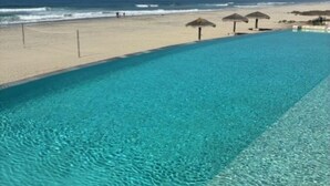 A clear blue pool adjacent to a sandy beach with a view of the ocean.
