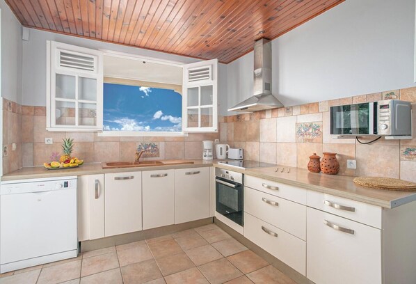 A kitchen with white cabinets, a wooden ceiling, a window with open shutters, a microwave, and a dishwasher. - Villa panoramic sea view & private pool (Le Robert)