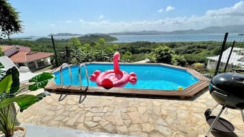 Ein Swimmingpool mit einem rosa Flamingo-Schwimmpflegegerät, umgeben von einer Steinterrasse, einem Grill und Blick auf das Meer und die Hügel.