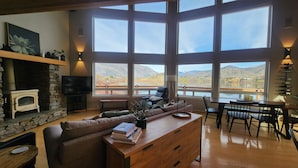 Ein geräumiges Wohnzimmer mit Kamin aus Stein, einer großen Couch, einem Essbereich und Blick auf Berge und See durch große Fenster.