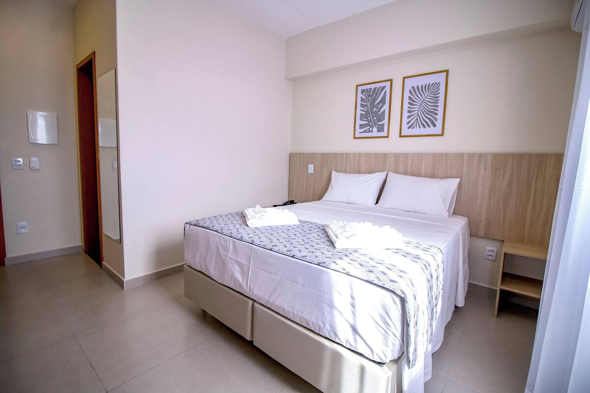 A hotel room with a large bed, a wooden headboard, two framed wall art pieces, a bedside table, and a power outlet.