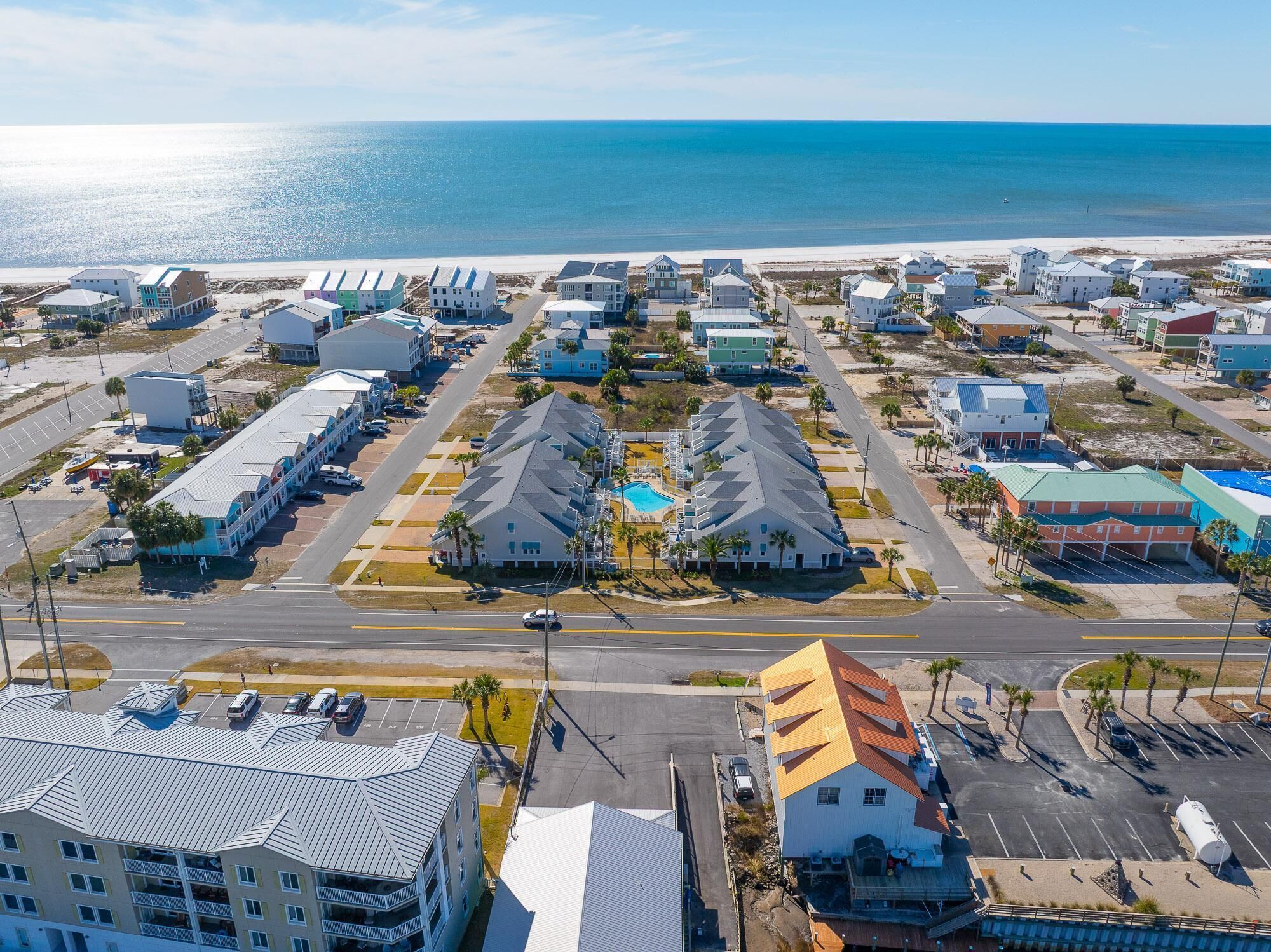 Vue aérienne d’une station balnéaire avec des propriétés situées en bord de mer, une piscine centrale et une route principale.