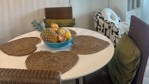 Un coin repas avec une table ronde, des sets de table en fibres naturelles, un saladier de fruits, des chaises en osier, un luminaire suspendu, un téléviseur fixé au mur et un buffet blanc aux portes en verre.