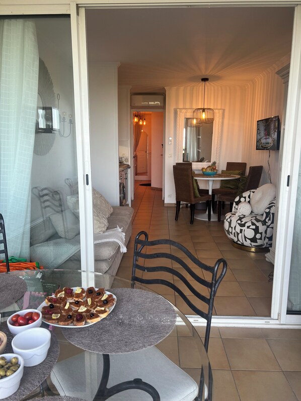 A dining area with a glass table, chairs, and a variety of food items. There is a sofa and a chair in the background, and a television mounted on the wall.