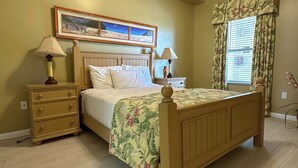 A bedroom with a wooden bed, two bedside tables with lamps, a nightstand, a window with floral curtains, and a painting above the bed. - Spacious 3-bedroom Beachfront Condo Pool, Large Balcony (Pensacola)