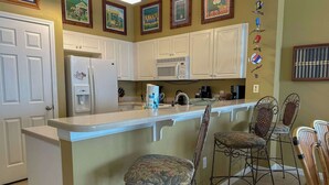 A kitchen with white cabinets, a refrigerator, a microwave, and a bar area with wicker chairs. - Spacious 3-bedroom Beachfront Condo Pool, Large Balcony (Pensacola)