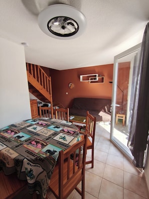 Un comedor con una mesa cubierta por un mantel estampado, sillas de madera, un ventilador de techo, una escalera y un puerta corrediza de vidrio que da a un balcón.